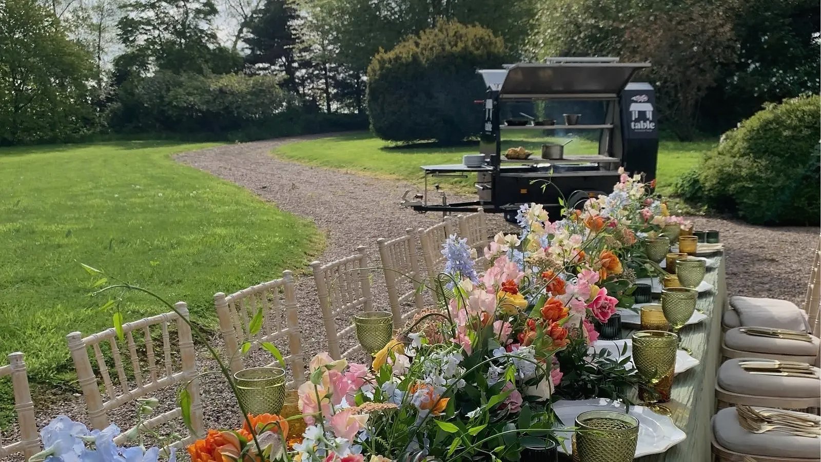 Flower-lined outdoor dining table with the Table Trailblazer charcoal BBQ behind it